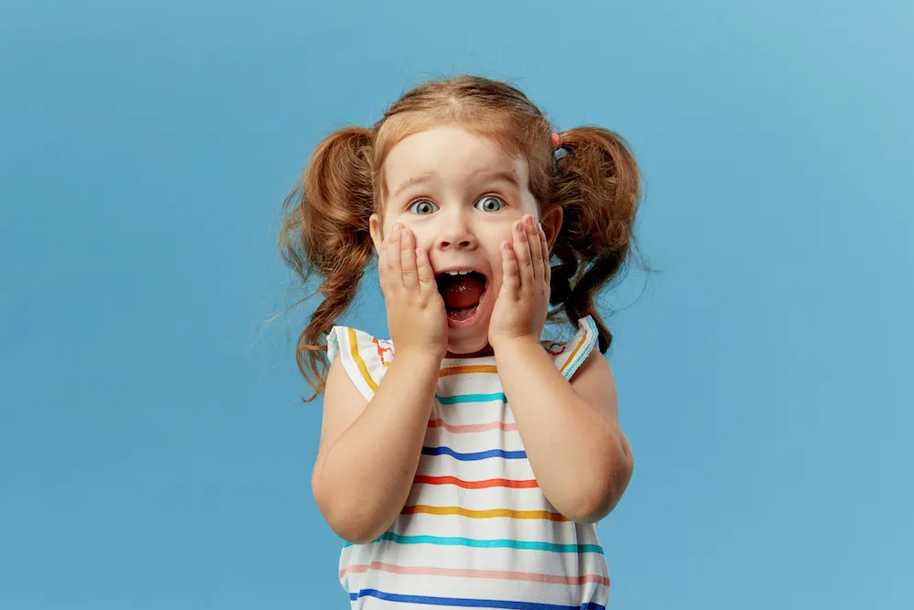 Portrait of surprised cute little toddler girl child standing isolated over blue background. Looking at camera. hands near open mouth
