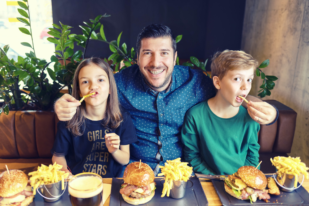 Modern dad with dental braces having fun with his kids eating hamburgers with french fries at trendy fast food restaurant